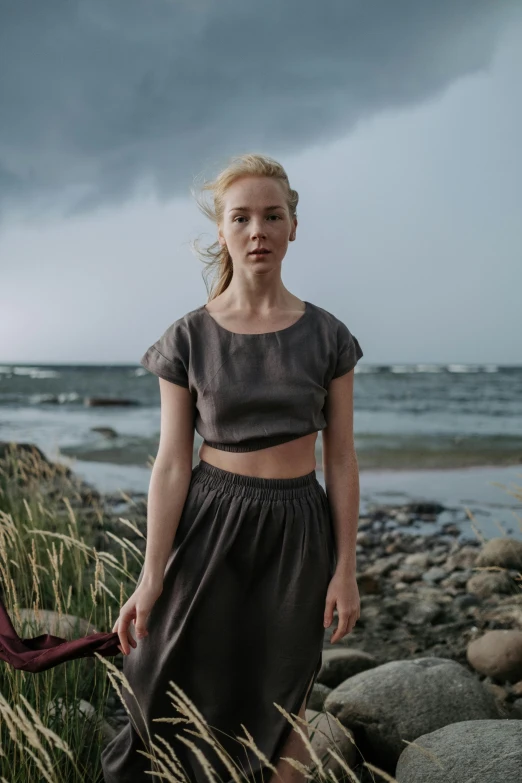 a woman standing on a rocky beach next to a body of water, inspired by Louisa Matthíasdóttir, renaissance, wearing a crop top, stormy skies, tv still, female outfit