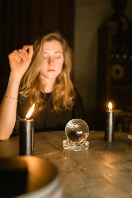 a woman sitting at a table with a crystal ball in front of her, inspired by Wilhelm Hammershøi, trending on pexels, candles in foreground, black orb of fire, vantablack, holding fire and electricity