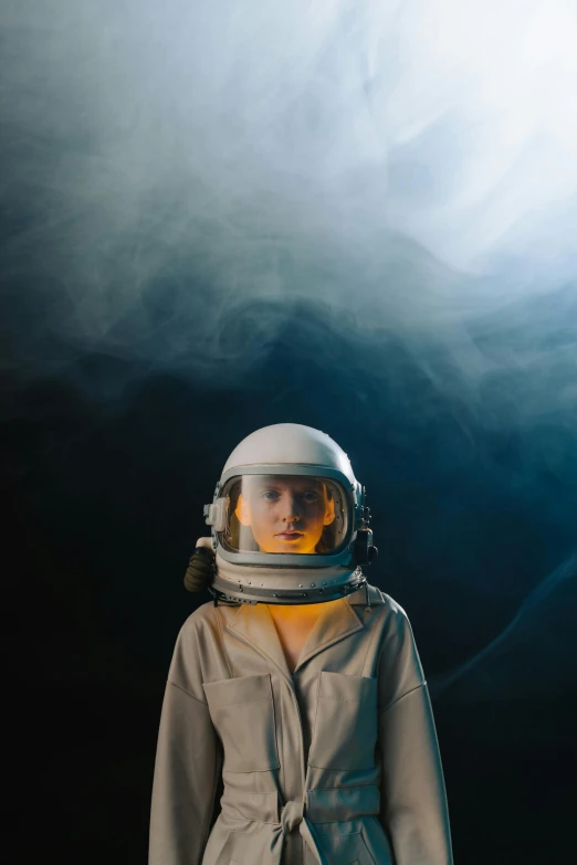 a woman in a space suit standing in front of a dark background, a portrait, pexels contest winner, light and space, in front of smoke behind, holding helmet, rex orange county, surrounded in clouds and light