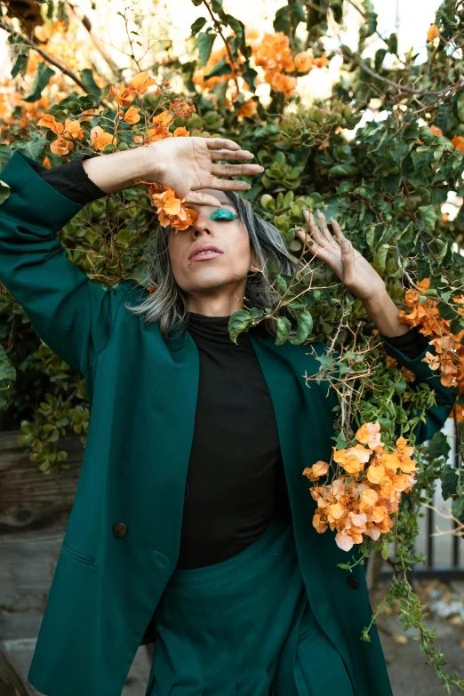 a woman standing in front of a bush of flowers, trending on pexels, color field, wearing green suit, black and orange coat, silver and muted colors, playful pose