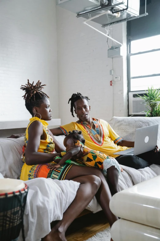 a couple of people sitting on a couch with a laptop, by Washington Allston, pexels contest winner, afrofuturism, yellow clothes, husband wife and son, wearing an african dress, nyc