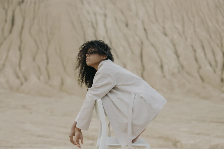 a woman sitting on a chair in the desert, pexels contest winner, afrofuturism, wearing lab coat and a blouse, curly haired, taupe, outlive streetwear collection