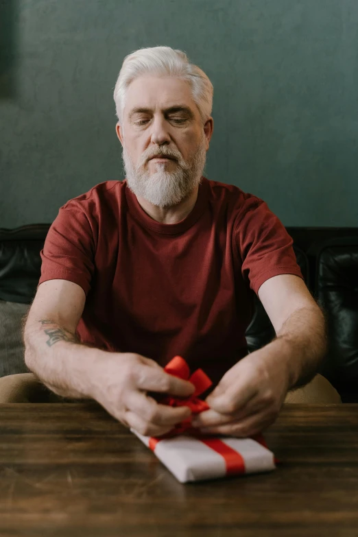 a man sitting at a table with a present, grey beard, red and grey only, puzzling, centered shot