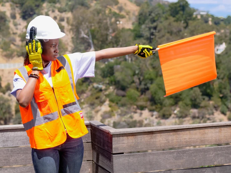 a woman in an orange safety vest holding an orange flag, unsplash, mulholland drive, guardrail, robotic extended arms, hillside