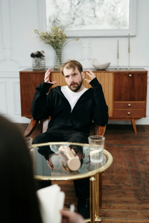 a man sitting in a chair in a living room, looking at each other mindlessly, with a beard and a black shirt, mr beast, shrugging