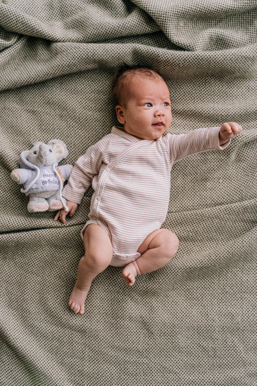 a baby laying on top of a blanket next to a stuffed animal, pexels contest winner, happening, muted colored bodysuit, full body frontal view, looking down on the camera, environmental shot