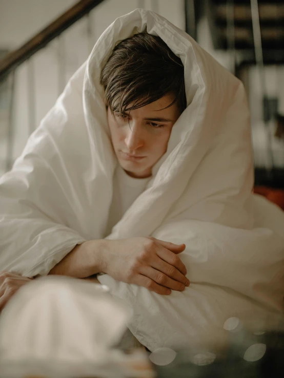 a man sitting on top of a bed under a blanket, trending on pexels, romanticism, boy with neutral face, wearing a white hospital gown, freezing, lgbtq