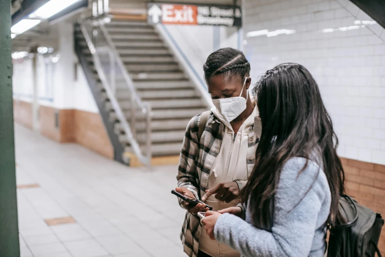 a couple of people standing on a subway platform, pexels contest winner, medical mask, woman holding another woman, ( ( dark skin ) ), modern technology