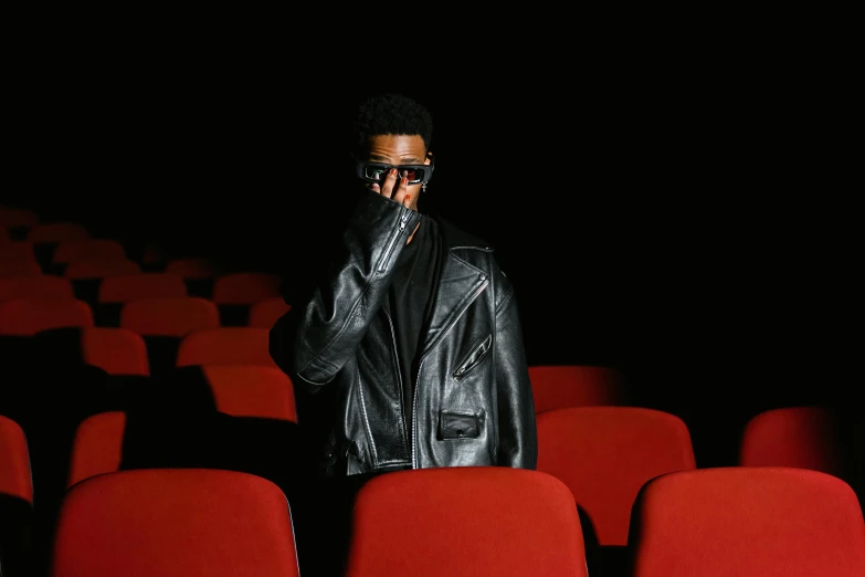 a man standing in front of a row of red chairs, an album cover, pexels, visual art, 2 1 savage, sitting in a movie theater, wearing black glasses, leather padding