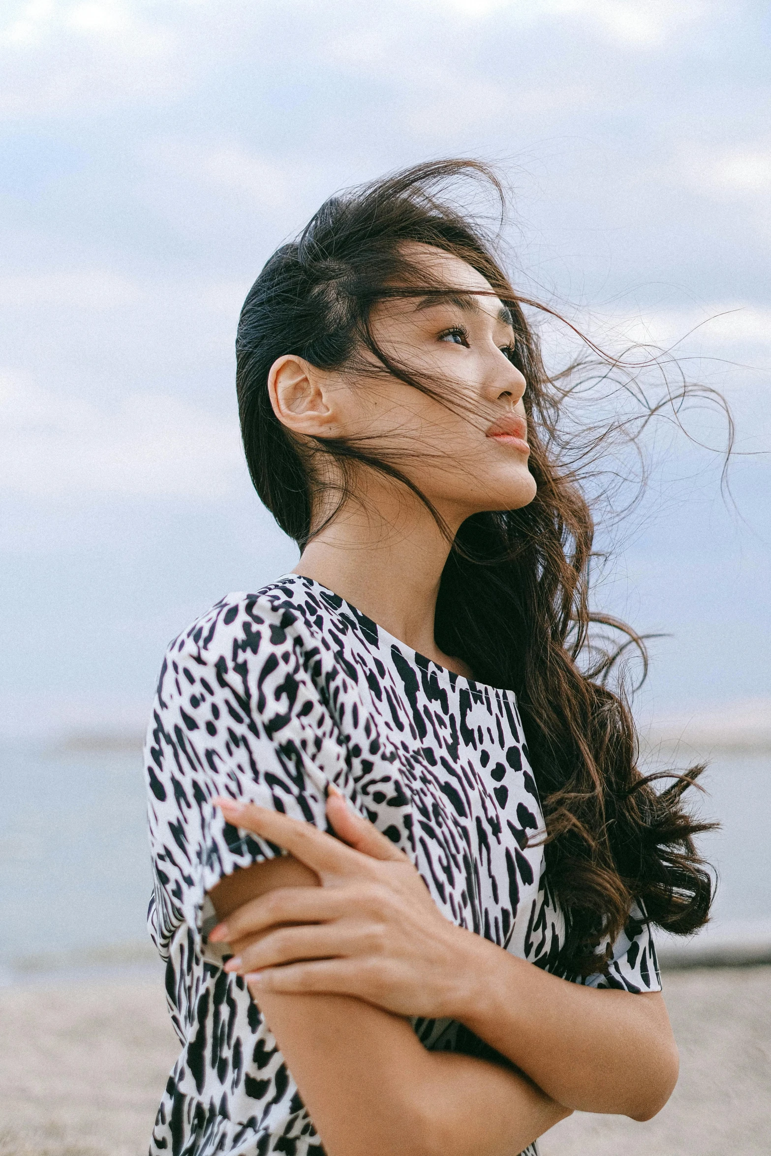 a woman standing on a beach next to the ocean, an album cover, inspired by Zheng Xie, trending on pexels, long windy hair style, patterned clothing, beautiful young asian woman, hands in her hair. side-view