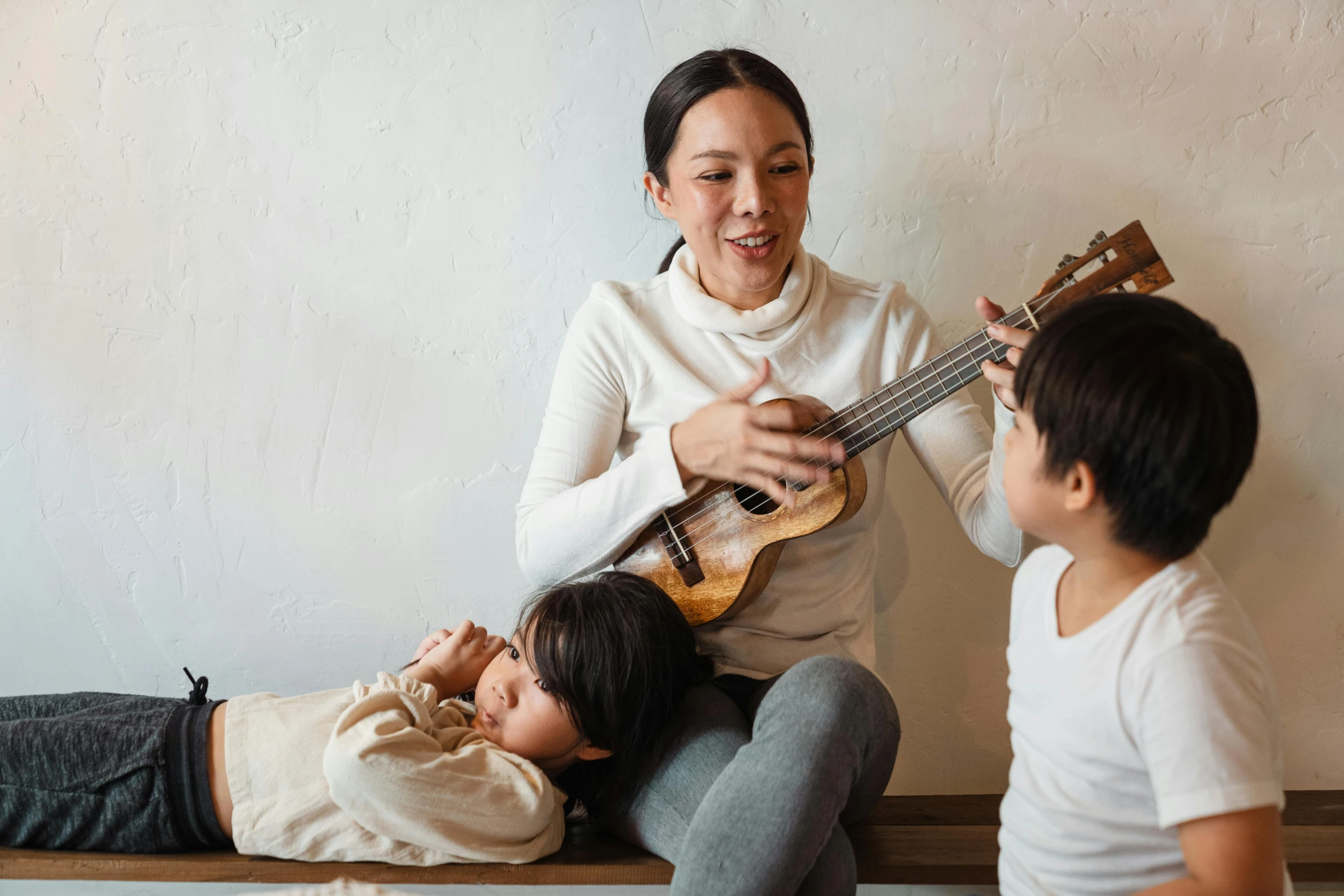 a woman sitting on top of a wooden bench next to a boy, an album cover, pexels contest winner, shin hanga, ukulele, families playing, ethnicity : japanese, at home