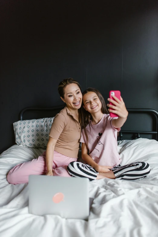 a couple of women sitting on top of a bed, by Julia Pishtar, pexels contest winner, taking a selfie, young asian girl, motherly, brown and pink color scheme