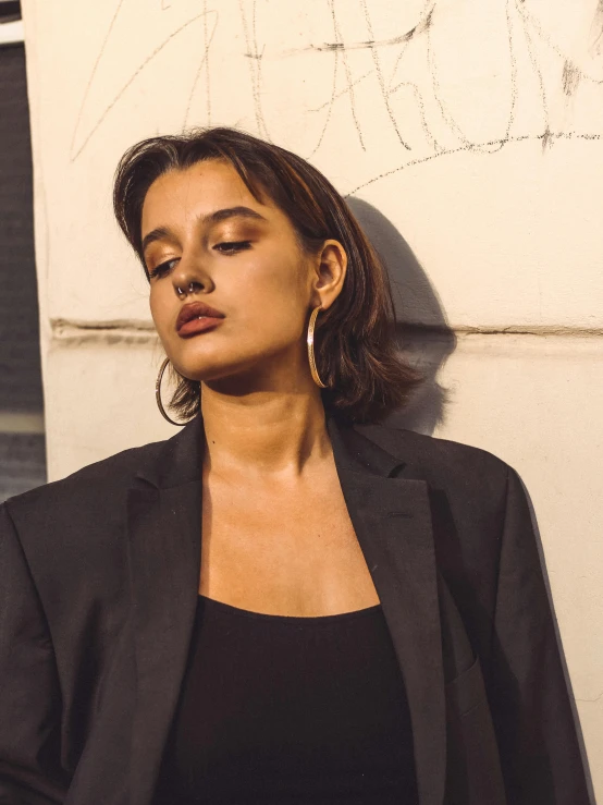 a woman leaning against a wall with her eyes closed, an album cover, by Emma Andijewska, trending on unsplash, photorealism, wearing a black blazer, hoop earrings, isabela moner, profile image