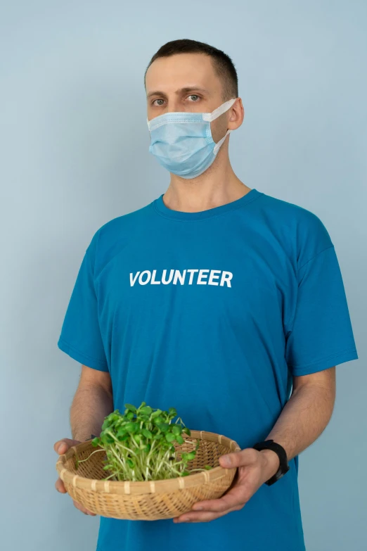 a man wearing a face mask holding a basket of plants, a picture, blue shirt, on a gray background, 2022 photograph, healthcare