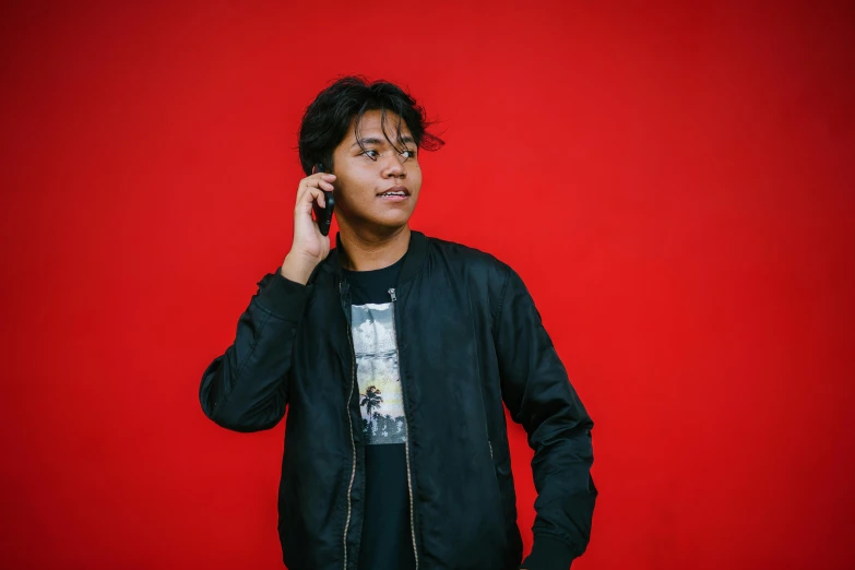 a man standing in front of a red wall talking on a cell phone, by Basuki Abdullah, wearing a black jacket, male teenager, plain background, caspar david