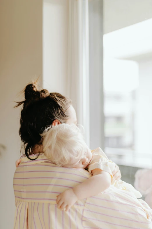 a woman holding a baby in her arms, pexels contest winner, looking out window, hugging each other, from back, on a pale background