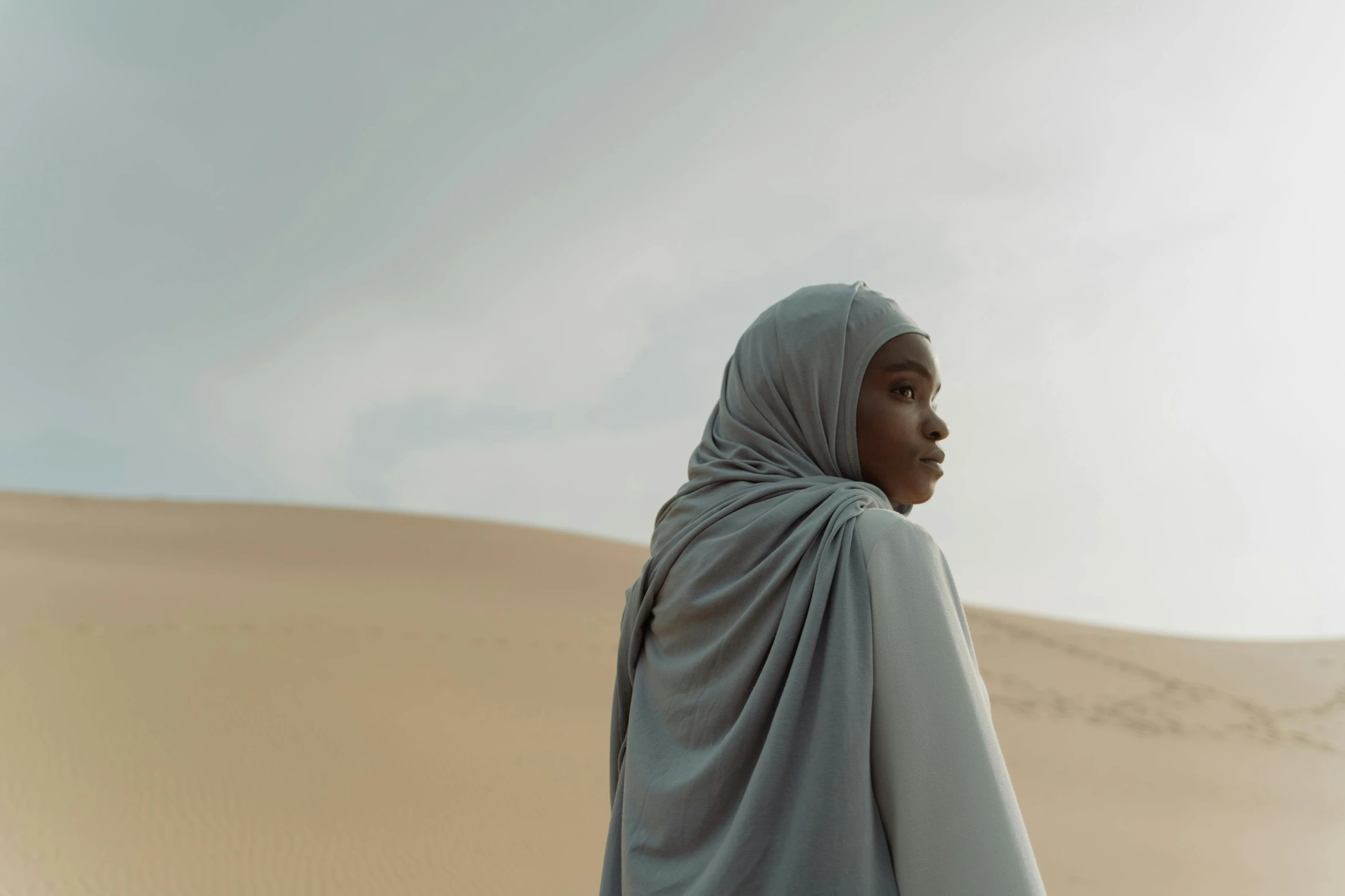 a woman standing in the middle of a desert, by Alasdair Grant Taylor, pexels contest winner, afrofuturism, wearing a grey robe, white hijab, still from l'estate, african american young woman