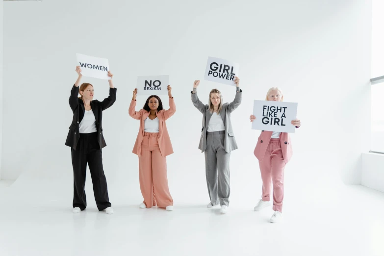 a group of women holding up signs in a white room, by Emma Andijewska, trending on pexels, feminist art, girl in a suit, on a gray background, full body with costume, modest