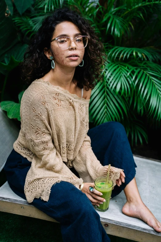 a woman sitting on a bench holding a drink, lush green, olive skinned, with glasses, wearing casual sweater