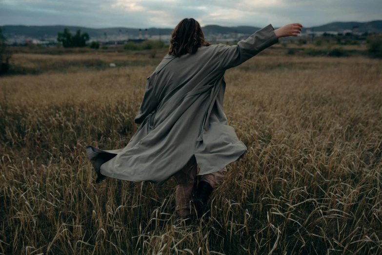 a woman standing in a field with her arms outstretched, an album cover, by Attila Meszlenyi, unsplash contest winner, renaissance, wearing a trenchcoat, movie still 8 k, windy hair, walking away