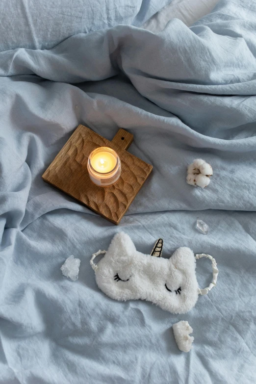 a white cat laying on top of a bed next to a lit candle, a picture, trending on pexels, soft blue texture, masks, unicorn horn, detailed product shot