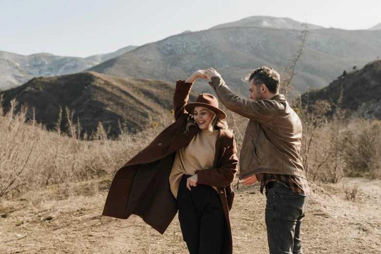 a man standing next to a woman on a dirt road, trending on pexels, romanticism, wearing a brown leather coat, girl dancing on cliff, smiling couple, thumbnail