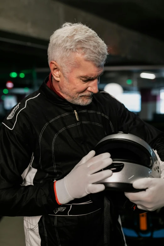 a man in a black jacket and white gloves holding a helmet, gray haired, formula 1 garage, body armour, carefully crafted