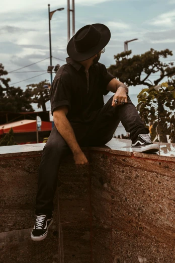 a man sitting on top of a stone wall, an album cover, trending on pexels, black stetson hat, rooftop party, fullbody photo, profile image