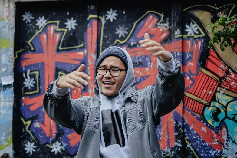 a man standing in front of a graffiti covered wall, an album cover, inspired by Gang Hui-an, pexels contest winner, he also wears a grey beanie, waving and smiling, performing a music video, te pae