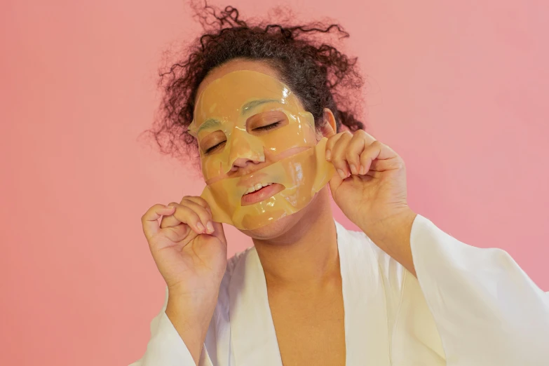 a woman putting a facial mask on her face, by Julia Pishtar, lime and gold hue, wearing translucent sheet, sassy pose, silicone skin