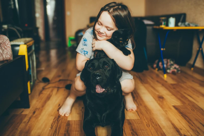 a little girl sitting on the floor with a black dog, pexels contest winner, cat and dog licking each other, tiny cat riding a labrador, hugging each other, thumbnail