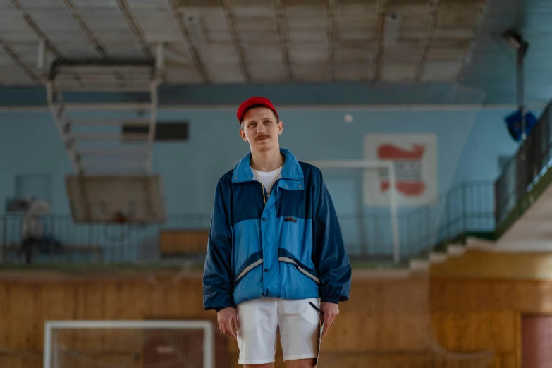 a man in a blue jacket and white shorts standing on a basketball court, by Attila Meszlenyi, unsplash, bauhaus, baggy clothing and hat, soviet style, indoor shot, portrait of small