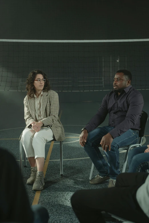 a group of people sitting in chairs in a room, looking at each other mindlessly, sports setting, video still, panel of black