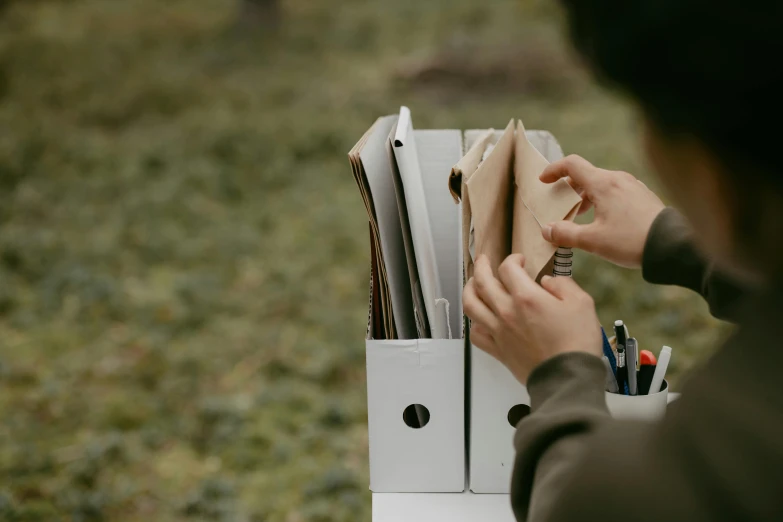 a person taking notes out of a white box, by Jessie Algie, pexels contest winner, forest picnic, holding books, cardboard, shot on 1 5 0 mm