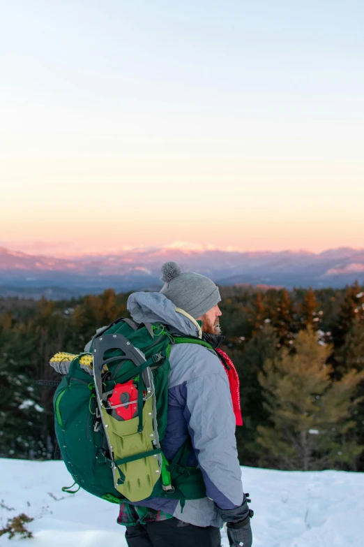 a man standing on top of a snow covered slope, by Jessie Algie, trending on unsplash, with a backpack, new hampshire, looking out at a sunset, in front of a forest background