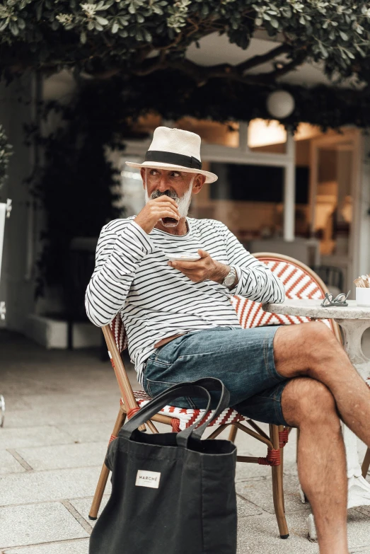 a man sitting on a chair eating a donut, white straw flat brimmed hat, wearing stripe shirt, grey beard, bra and shorts streetwear