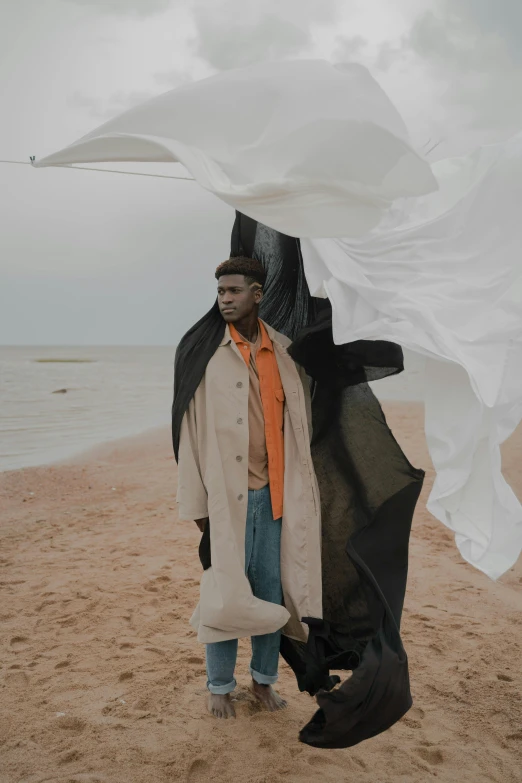 a man standing on top of a sandy beach, an album cover, by Attila Meszlenyi, trending on unsplash, surrealism, he is wearing a trenchcoat, draped in flowing fabric, heron preston, model posing