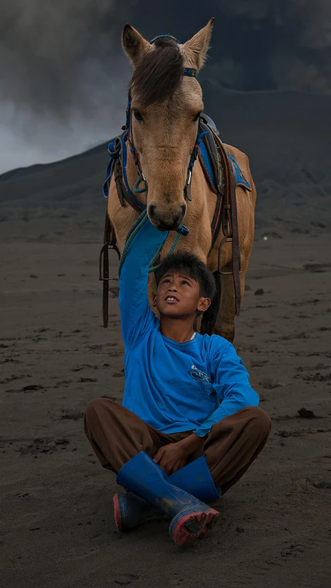 a man sitting on the ground next to a horse, inspired by Steve McCurry, pexels contest winner, sumatraism, cute boy, black volcano afar, portrait of tall, blue