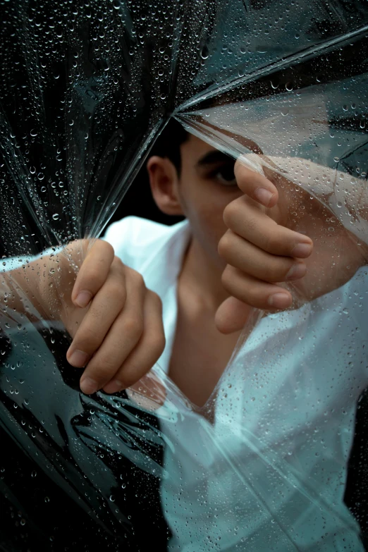 a man holding an umbrella in the rain, an album cover, by irakli nadar, hands shielding face, close - up photograph, male teenager, wearing translucent sheet