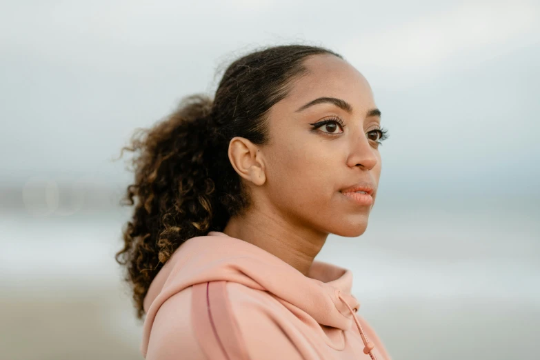 a woman standing on top of a beach next to the ocean, by Lily Delissa Joseph, pexels contest winner, renaissance, closeup headshot portrait, ashteroth, wearing a pastel pink hoodie, portrait of vanessa morgan