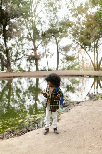 a little girl standing next to a body of water, walking at the park, looking at his phone, wearing adventuring gear, off - white collection