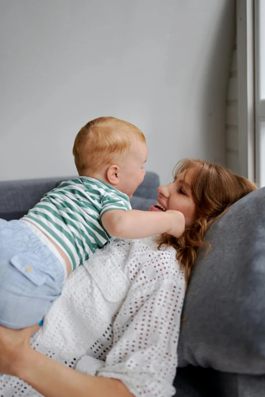 a woman laying on a couch holding a baby, pexels contest winner, happening, brunette boy and redhead boy, up to the elbow, playing, on grey background