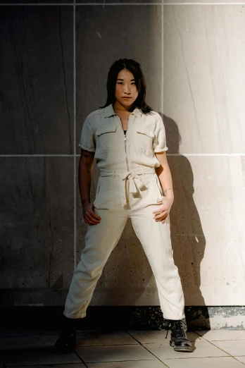 a woman in a white jumpsuit standing in front of a wall, an album cover, inspired by Ruth Jên, unsplash, wearing cargo pants, asian woman, standing in a dimly lit room, wearing track and field suit