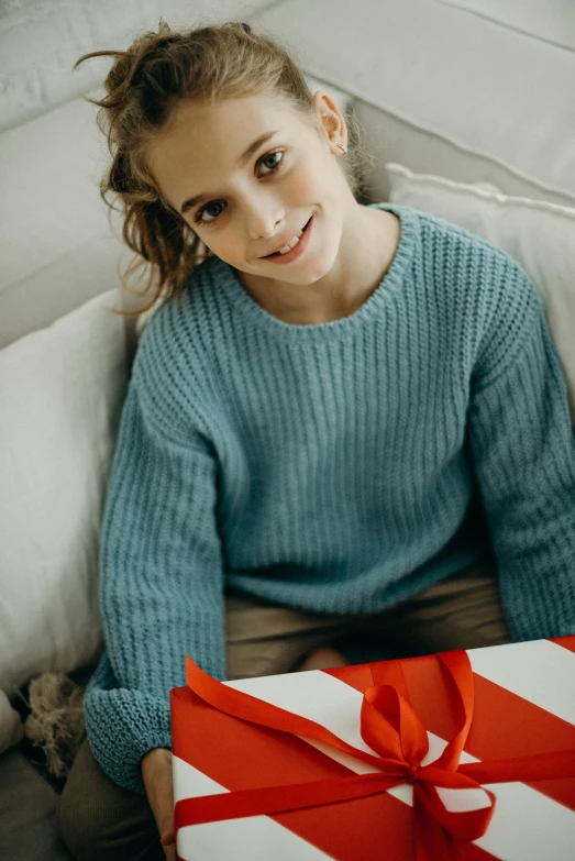 a young girl sitting on a couch holding a present, pexels contest winner, blue sweater, light blue, product introduction photo, subtle smile