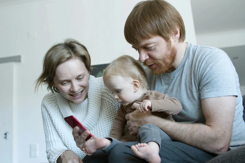 a man and woman sitting on a couch with a baby, a photo, by Eero Järnefelt, pexels, realism, holding a very advance phone, square, educational, brown