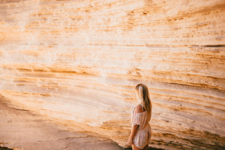 a woman standing in front of a rock wall, by Lee Loughridge, pexels contest winner, minimalism, sandy colours, amber glow, sandstone, showing her shoulder from back