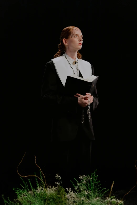 a woman standing on a stage holding a book, an album cover, inspired by Rogier van der Weyden, unsplash, wearing a black noble suit, white on black, sophia lillis, holy ceremony