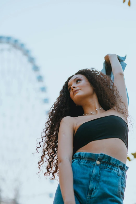 a woman standing in front of a ferris wheel, wearing a crop top, long wild black curly hair, hands in her hair, 2019 trending photo