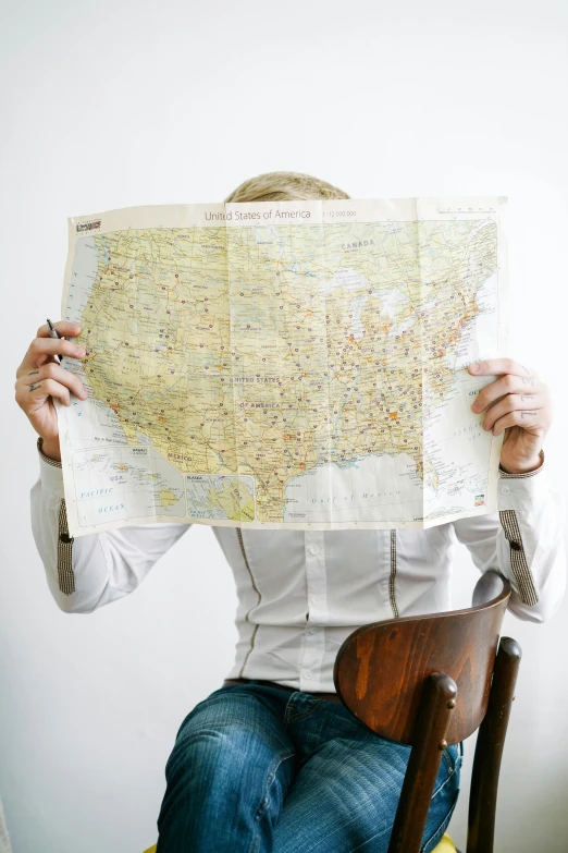 a man sitting on a chair holding up a map, by Jessie Algie, trending on unsplash, regionalism, american total portrait, 15081959 21121991 01012000 4k, panel, conde nast traveler photo