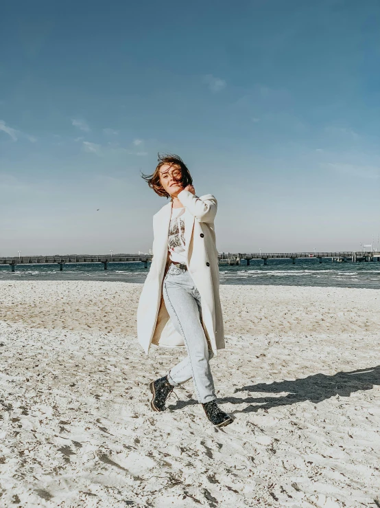 a woman standing on top of a sandy beach, wearing a white winter coat, happy vibes, doing a sassy pose, berlin fashion